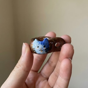 May include: A brown ceramic ring with a painted cat face in blue and white. The ring is held in a hand, showcasing the handmade details and unique design. The cat's face has a blue and white color scheme.
