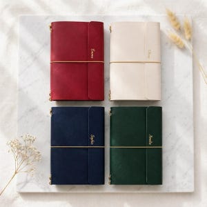 May include: Four leather journals in red, cream, navy, and forest green. Each journal has a gold elastic closure and the name "Emma", "Olivia", "Sophia", or "Amelia" in gold lettering. The journals are arranged on a white marble surface.