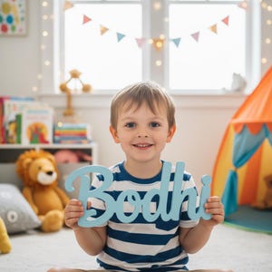 May include: A child holds a light blue wooden sign with the name "Bodhi" in a cursive font. The child is wearing a blue and white striped top and smiling. The background shows a playroom with a tent and soft toys.
