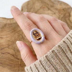 May include: A gold-toned ring featuring a light purple oval-shaped centerpiece. The centerpiece has a detailed brown and beige design. The ring is worn on a finger, showcasing the intricate details and craftsmanship of the jewelry.