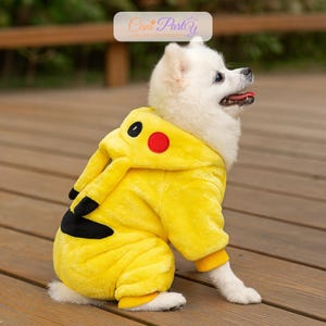 May include: A small white dog wearing a yellow Pikachu costume with black and red accents. The dog is sitting on a wooden surface. The costume has a hood with ears and a tail.