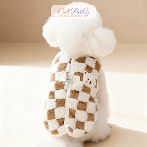 May include: A dog wearing a brown and white checkered vest with a zipper closure. The vest has a small bear applique on the front. The text "Cani Party - DOG AND CAT COSTUMES" is in the upper right corner.