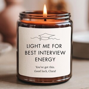 May include: A lit candle in a glass jar with a white label. The label reads "LIGHT ME FOR BEST INTERVIEW ENERGY" with a graduation cap graphic. The text "You've got this. Good luck, Clara!" is also on the label. The candle sits on a wooden surface.