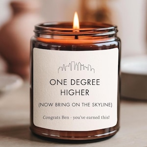 May include: A lit candle in a glass jar with a label that reads "ONE DEGREE HIGHER" and "(NOW BRING ON THE SKYLINE)". The label also includes a skyline graphic and the text "Congrats Ben - you've earned this!"