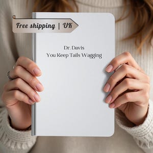 May include: A white notebook with the text "Dr. Davis You Keep Tails Wagging" is held by a person. The notebook has a silver edge and is blank except for the text. The person is wearing a cream-colored sweater.