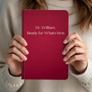 Könnte beinhalten: Ein weinrotes Notizbuch mit dem weißen Text "Dr. William, Ready for What's Next". Das Notizbuch wird von einer Person in einem cremefarbenen Pullover gehalten.