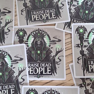 May include: Square stickers with a skeletal figure in a hooded robe and crown, holding a staff. The text "I RAISE DEAD PEOPLE" is displayed. The design is in black, green, and grey.