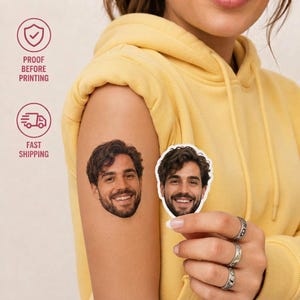 Puede incluir: Un tatuaje temporal personalizado que presenta un retrato realista de un hombre con cabello castaño oscuro y barba, aplicado en un brazo. Una pegatina troquelada a juego del mismo retrato se sostiene entre los dedos, mostrando los rasgos faciales.