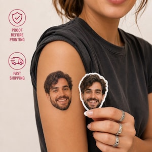 Può includere: Un tatuaggio temporaneo personalizzato che mostra il ritratto fotografico di un uomo sorridente con capelli scuri ondulati e barba. Il tatuaggio è applicato sulla pelle e una versione adesiva identica viene tenuta vicino.