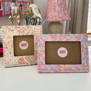 Pu&ograve; includere: Due cornici decorative con motivi floreali e a stella. Una cornice ha un design floreale rosa, giallo e blu, mentre l'altra presenta un motivo a stella. Entrambe le cornici hanno un interno marrone e un adesivo "SAINT MALO".