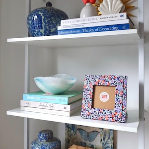 Pu&ograve; includere: Una mensola bianca espone oggetti per la casa. Una cornice floreale con un adesivo Saint Malo, libri, una ciotola blu e bianca e un vaso di ceramica blu e bianco sono visibili. Libri: "Domino" e "beach life."