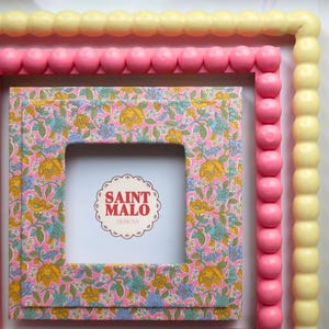 Pu&ograve; includere: Una cornice quadrata con un motivo floreale rosa, giallo e blu. La cornice ha un centro bianco con il testo "SAINT MALO DESIGNS". La cornice &egrave; circondata da un bordo di perline rosa e giallo.