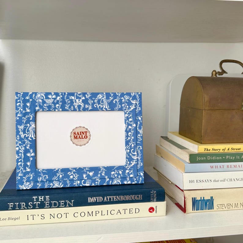 Pu&ograve; includere: Una cornice blu con un motivo floreale bianco e un interno bianco, appoggiata su libri. La cornice ha un adesivo che dice "SAINT MALO". Un piccolo forziere in ottone e altri libri sono a destra.