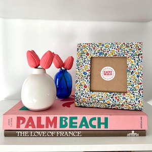 Pu&ograve; includere: Una cornice quadrata a fantasia floreale con interno marrone, accanto a un vaso bianco con tulipani rosa e un vaso blu. La cornice ha un adesivo con la scritta "SAINT MALO". La cornice e i vasi sono sopra due libri, uno rosa e uno marrone.