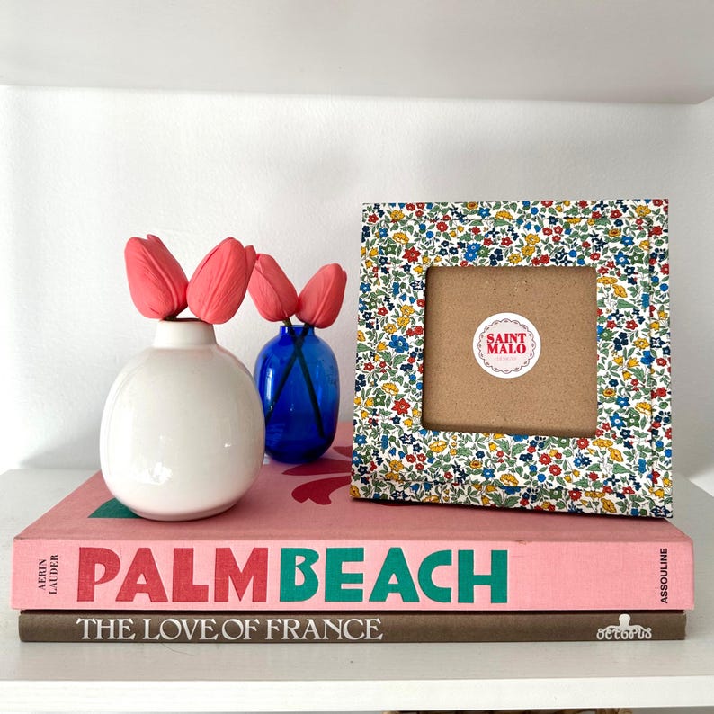Pu&ograve; includere: Una cornice quadrata a fantasia floreale con interno marrone, accanto a un vaso bianco con tulipani rosa e un vaso blu. La cornice ha un adesivo con la scritta "SAINT MALO". La cornice e i vasi sono sopra due libri, uno rosa e uno marrone.