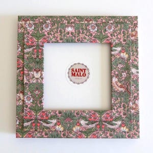 Pu&ograve; includere: Cornice quadrata con un motivo floreale e di uccelli in rosa, verde e bianco. La cornice ha un interno bianco e un adesivo con la scritta "SAINT MALO DESIGNS".