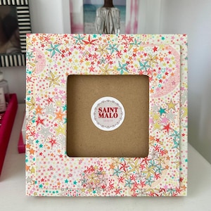 Pu&ograve; includere: Cornice quadrata con un colorato motivo a stelle in rosso, giallo, blu e rosa. La cornice ha un'apertura quadrata e un retro marrone. Un adesivo circolare con la scritta "SAINT MALO DESIGNS" &egrave; visibile.