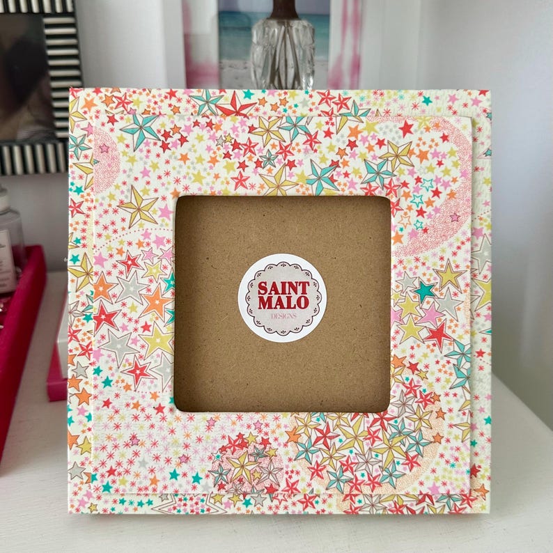 Pu&ograve; includere: Cornice quadrata con un colorato motivo a stelle in rosso, giallo, blu e rosa. La cornice ha un'apertura quadrata e un retro marrone. Un adesivo circolare con la scritta "SAINT MALO DESIGNS" &egrave; visibile.