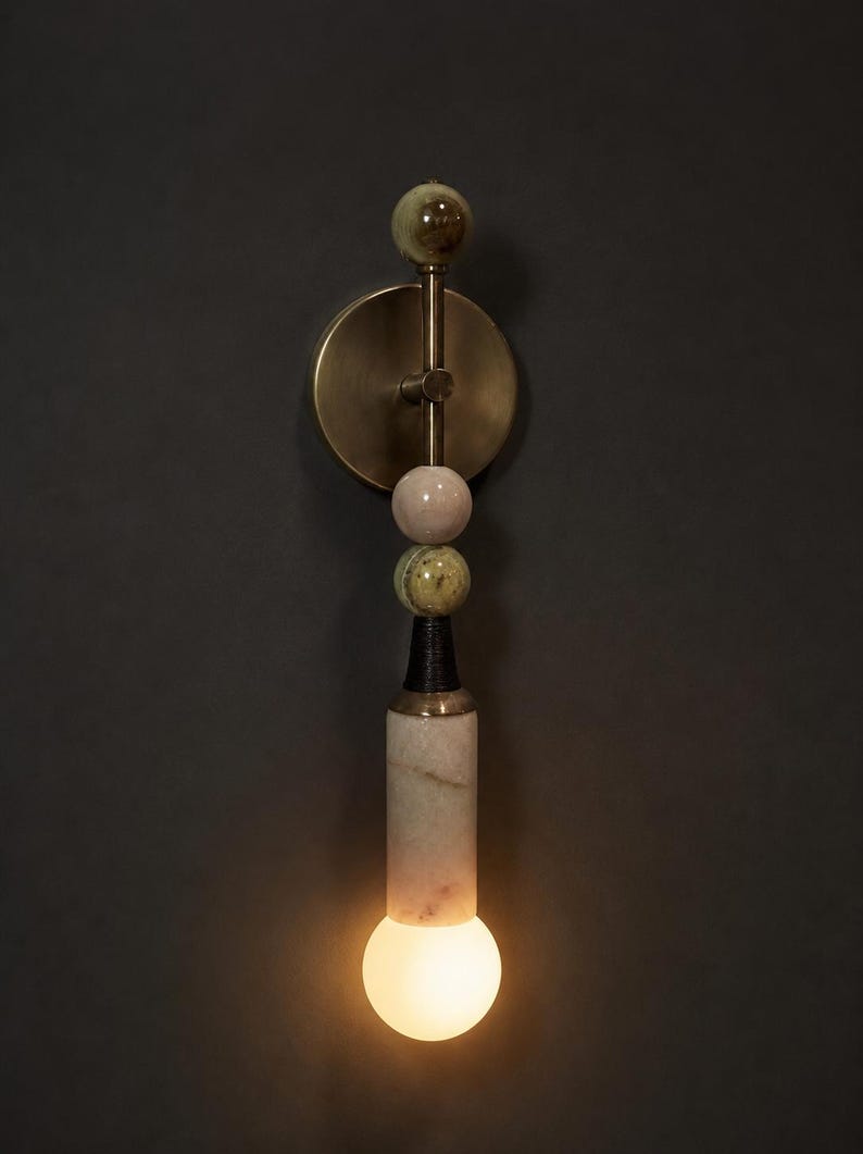 May include: A modern wall sconce with a brass finish and a series of decorative spheres in shades of green and pink. The sconce features a cylindrical base with a marble-like pattern and a glowing bulb at the bottom.