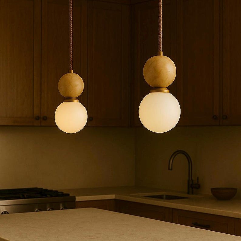 May include: Two pendant lights with a wooden sphere above a white globe. The lights hang from brown cords against a dark wood background. The kitchen setting includes a countertop, sink, and cabinets.