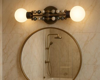 May include: A decorative bathroom light fixture with two round, glowing bulbs. The fixture has a dark bronze finish and is adorned with beads in shades of white, gray, and beige. A round mirror with a gold frame is below the light.