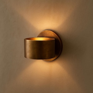 Könnte beinhalten: Eine wandmontierte, zylindrische Wandleuchte mit warmem Messing-Finish. Die Leuchte hat eine kreisförmige Rückplatte und einen kurzen, oben offenen Zylinder, der eine Lichtquelle enthält. Das Licht wirft einen sanften Schein auf die Wand und erzeugt eine warme Atmosphäre.
