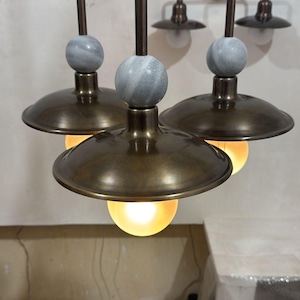May include: Three pendant lights with bronze-colored shades and stems, each topped with a gray marble sphere. The lights hang from the ceiling, with a matching two-light sconce on the wall in the background. The light bulbs are glowing.