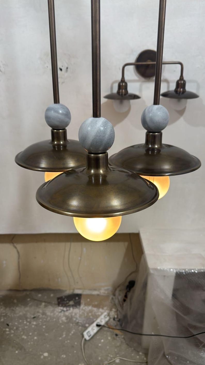 May include: Three pendant lights with bronze-colored shades and stems, each topped with a gray marble sphere. The lights hang from the ceiling, with a matching two-light sconce on the wall in the background. The light bulbs are glowing.