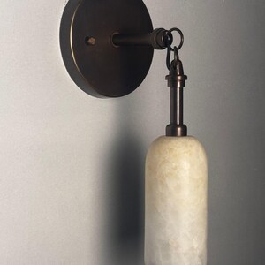 May include: A wall-mounted light fixture featuring a circular dark bronze base and a matching metal arm. A cylindrical, translucent, off-white stone shade hangs vertically from the arm, secured by a small metal ring and a short, slender stem.