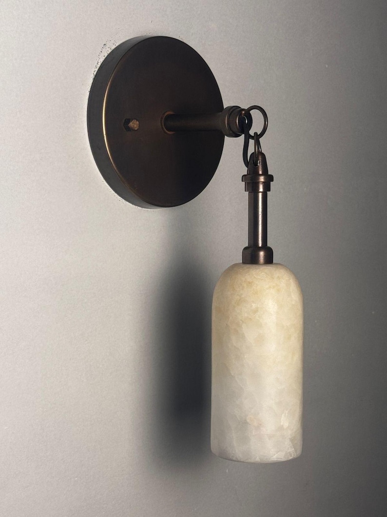 May include: A wall-mounted light fixture featuring a circular dark bronze base and a matching metal arm. A cylindrical, translucent, off-white stone shade hangs vertically from the arm, secured by a small metal ring and a short, slender stem.