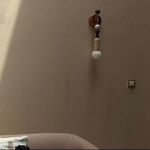 May include: A modern wall sconce with a gold-toned circular base and a series of decorative elements. The sconce features a light bulb at the bottom, with a wooden and black accent. The wall is a neutral beige color.