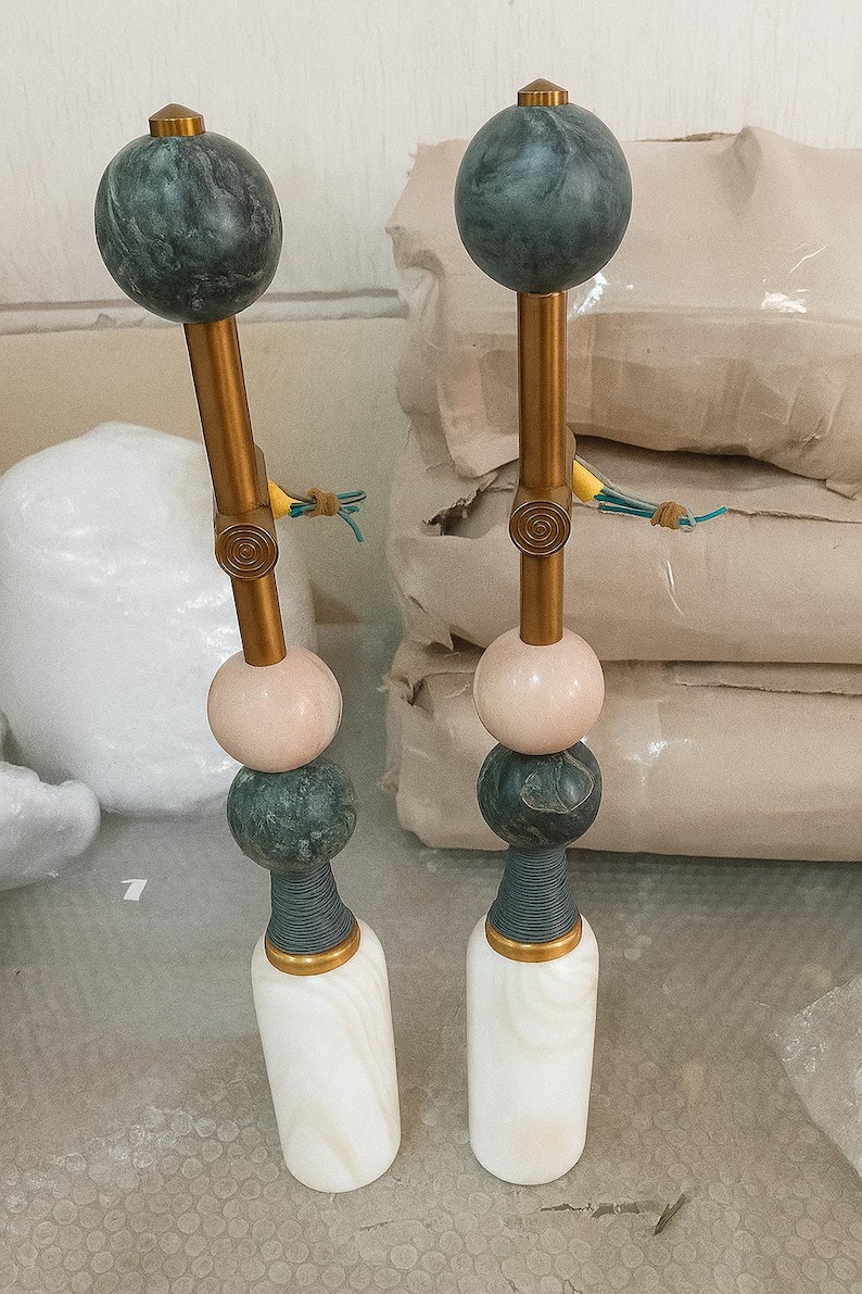 May include: Two decorative objects with a gold-colored stem, topped with a dark green sphere. The stem has a pink sphere, a dark green sphere, and a white base. The objects are likely decorative items or sculptures.