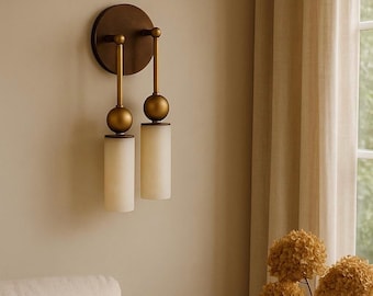 May include: A wall-mounted light fixture with a circular bronze backplate and two arms. Each arm has a gold sphere and a cylindrical, off-white glass shade. The fixture is mounted on a beige wall, with a neutral-toned curtain and dried flowers in the background.