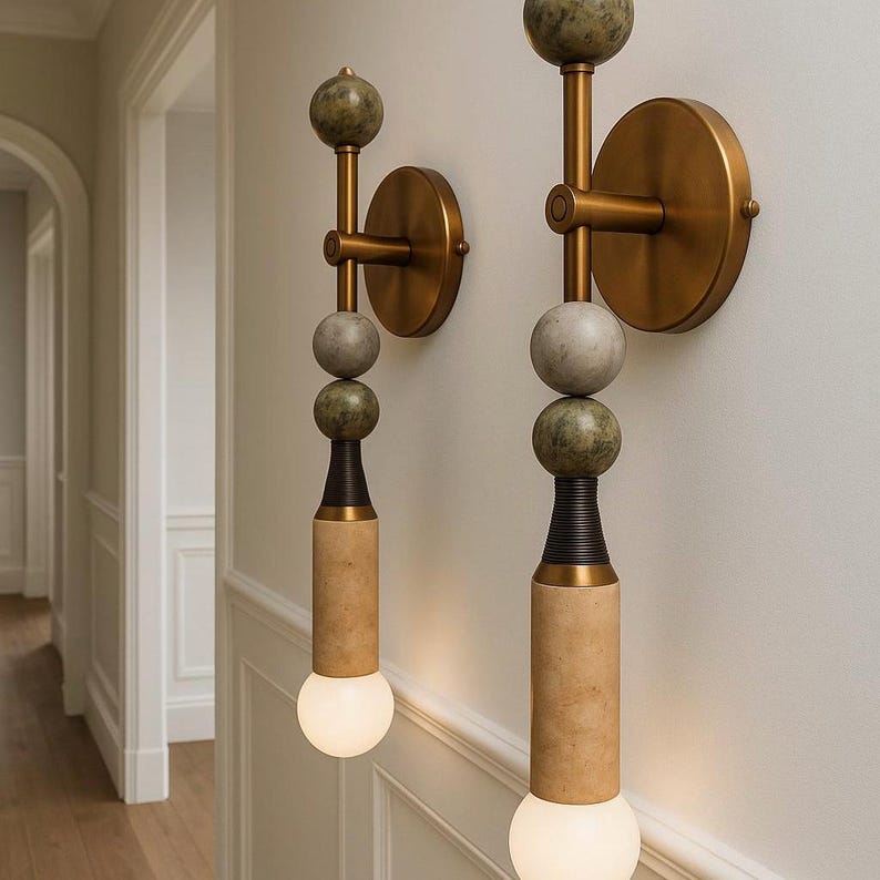 May include: Two wall sconces with a modern design. Each sconce features a brass-colored circular base, a series of stone spheres in shades of green and gray, and a cylindrical base with a warm, textured finish. The sconces have a warm, glowing light.