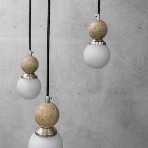 May include: Three pendant lights with a modern design. Each light features a spherical, frosted glass bulb, topped with a textured, beige sphere and a silver-toned metal fixture. Black cords suspend the lights against a gray background.
