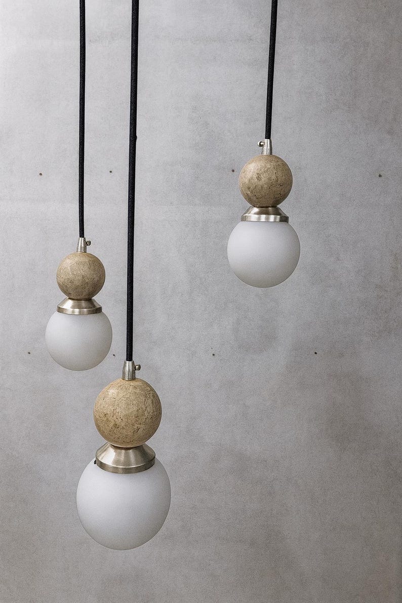 May include: Three pendant lights with a modern design. Each light features a spherical, frosted glass bulb, topped with a textured, beige sphere and a silver-toned metal fixture. Black cords suspend the lights against a gray background.