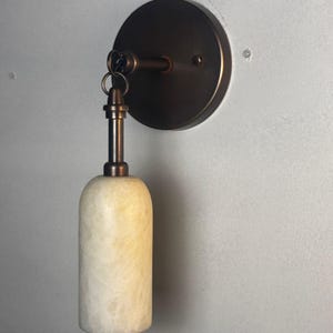 May include: A wall-mounted light fixture with a circular bronze backplate and a cylindrical, off-white alabaster shade. The fixture has a bronze arm and a small ring connecting the shade to the arm. The light is designed for indoor use.