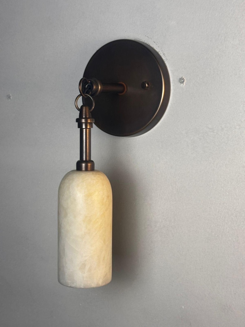May include: A wall-mounted light fixture with a circular bronze backplate and a cylindrical, off-white alabaster shade. The fixture has a bronze arm and a small ring connecting the shade to the arm. The light is designed for indoor use.