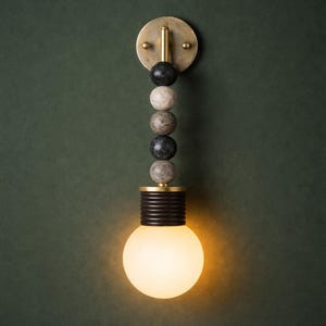 May include: A modern wall sconce with a round, brass-colored mounting plate and a series of decorative stone beads. The light fixture features a dark brown, cylindrical base and a large, round, frosted glass bulb. The sconce is mounted on a dark green wall.