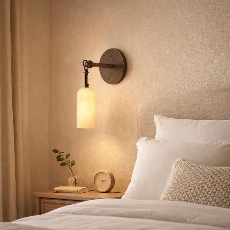 May include: A wall-mounted light fixture with a round, bronze-colored backplate and a curved arm. The light has a cylindrical, off-white glass shade. The fixture is mounted on a beige wall above a bed with white pillows.