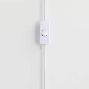 Op de afbeelding: Een witte inline aan/uit-schakelaar met een afgeronde rechthoekige vorm, verbonden met twee witte elektrische snoeren. De schakelaar heeft een enkele, ovaalvormige knop in het midden. De achtergrond is een effen witte muur.