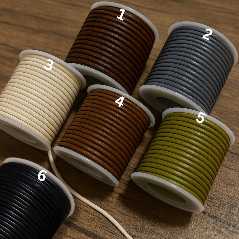 May include: A collection of six spools of leather cord in various colors, including brown, gray, cream, tan, olive green, and black. Each spool is neatly wound and labeled with a number. The cords are displayed on a wooden surface.