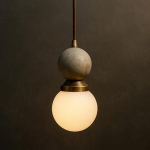 May include: A modern pendant light with a spherical design. It features a frosted glass globe at the bottom, a brass-colored metal accent, and a textured gray sphere above. The light hangs from a brown cord against a dark background.
