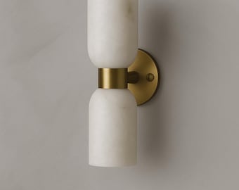 May include: A modern wall sconce featuring a white marble design with a brass accent. The light fixture has a cylindrical shape with a rounded top and bottom, connected by a brass band. The sconce is mounted on a round brass backplate.