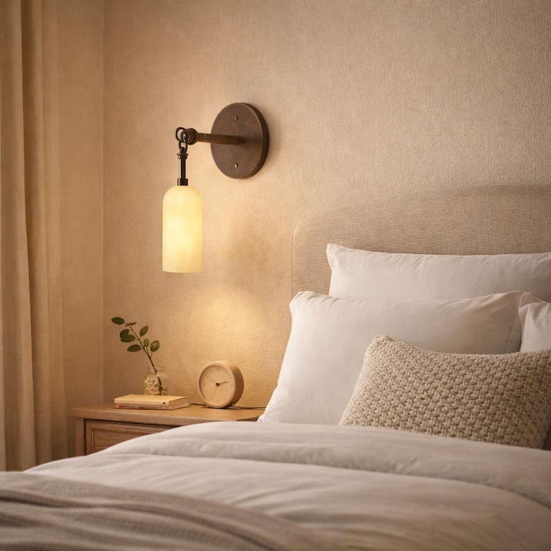 May include: A wall-mounted light fixture featuring a circular dark metal base and a cylindrical, frosted glass shade that emits a warm glow. The lamp is positioned above a wooden nightstand next to a bed with white pillows.