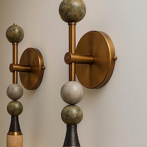 May include: Two wall sconces with a modern design. Each sconce features a brass-colored circular base and arm, with stacked spheres in shades of green and gray. The base is a light tan color with a dark brown accent.