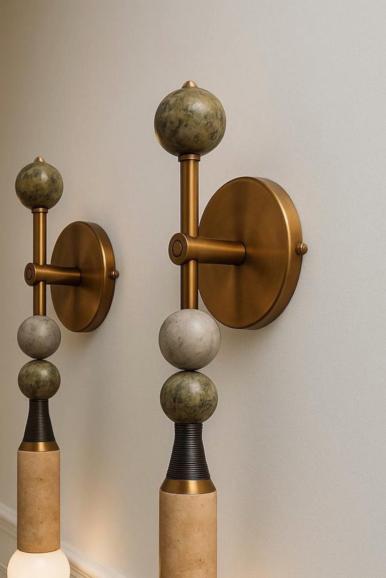 May include: Two wall sconces with a modern design. Each sconce features a brass-colored circular base and arm, with stacked spheres in shades of green and gray. The base is a light tan color with a dark brown accent.