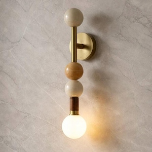 May include: A contemporary wall sconce with a brass finish and a series of spherical accents. The sconce features a warm, glowing bulb at the bottom, with a light beige, brown, and white colour scheme. The fixture is mounted on a circular brass base.