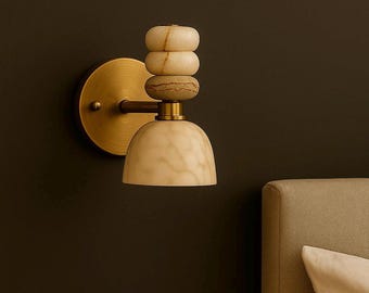 May include: A wall-mounted light fixture featuring a circular brass backplate and a horizontal arm. The lamp includes a stack of three rounded, textured stone elements in cream and beige tones, topped by a dome-shaped, marbled stone lampshade.