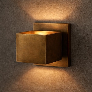 Op de afbeelding: Een vierkante wandlamp met een metallic bronzen afwerking. Het armatuur heeft een doosachtig ontwerp gemonteerd op een platte vierkante achterplaat, die een warme, gerichte lichtgloed uitstraalt vanuit zowel de boven- als onderopeningen.